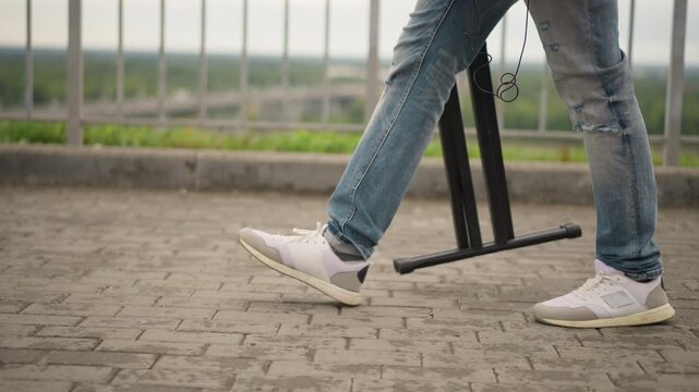 street musician walking with tripod sidewalk denim jeans white sneakers carrying mic stand between sets, overcast urban railing background, slow stride capturing departure after outdoor gig, ambient