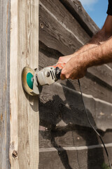 A man with a powerful tool with an angle grinder cleans the wood of the fence - grinding the boards with a coarse grinder brush