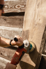 A man with an electric grinder in his hands sands the wooden surface of a fence - cleaning wood before renovation and repair - applying a protective compound