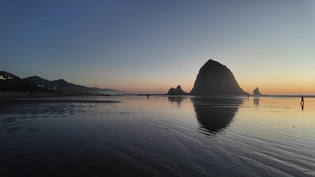 4K sunset at Cannon Beach, Oregon with people on the shoreline, Haystack Rock, calm Pacific Ocean waves, and golden reflections on wet sand. Travel and tourism footage