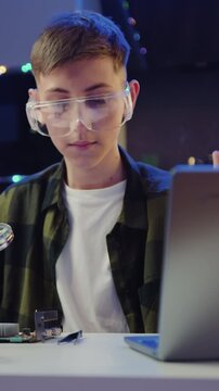 Boy in protective glasses soldering panel using soldering iron during revision training video on laptop in the evening at home
