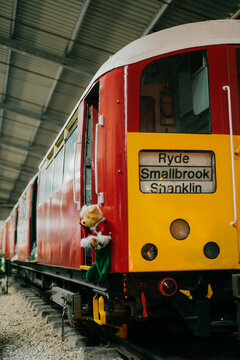 A train with a teddy bear on it is parked in a train station. The train is yellow and red and has the name Rylee Smallbrook Shanklin on the side
