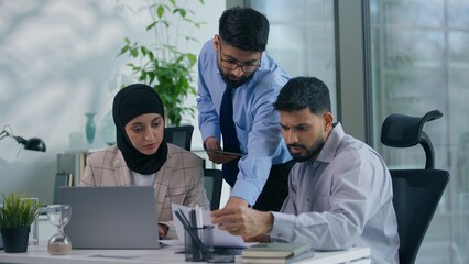 Business Arabian team three Indian Muslim colleagues in office discuss work project with documents looking at laptop using digital tablet working process co-workers analyze financial data at table