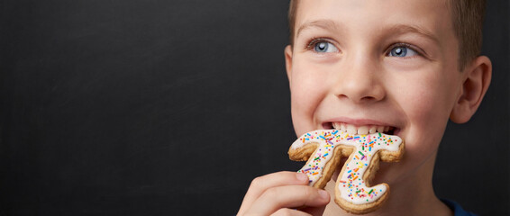 Infant nibbles on festive pastry, chalkboard backdrop, upward gaze, sprinkled confection, joyful expression. Academic setting, playful learning. International Day of Mathematics