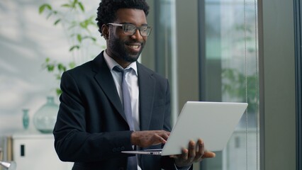 African American businessman male business man in office looking out window typing laptop smiling happy working at workplace using corporate software chatting internet browsing analyzing online data