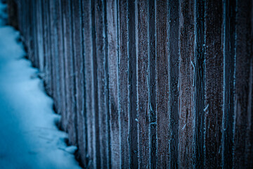 Close-up of a vertical wooden fence covered in frost, with snow on the ground and a gloomy blue winter atmosphere.