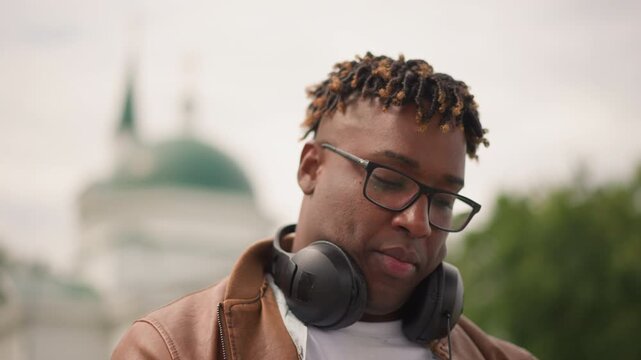 black man with headphones contemplative portrait, soft cinematic light, leather jacket, city dome background, shallow depth, slow motion closeups, thoughtful expression, urban calm mood, street - Powered by Adobe