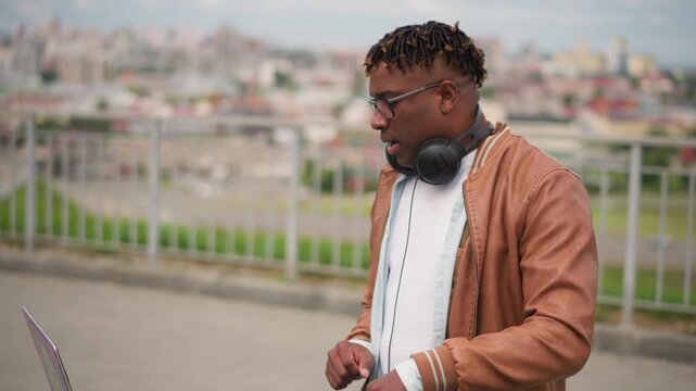 black producer adjusting headphones near laptop, closeup portraits with brown jacket and headphones around neck, focused expression, laptop and controller on stand, skyline blurred, cityscape