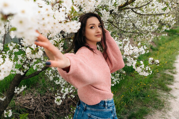 Fashionable young woman gently touching cherry blossoms while posing in a blooming spring orchard, enjoying the beauty of nature and the pleasant weather