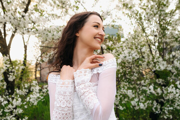 Romantic brunette woman wearing white lace dress is posing in blossoming garden, enjoying spring warm weather, smiling and touching her shoulders