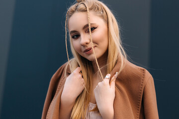 Fashionable influencer with long blonde braids wearing a stylish brown coat posing outdoors against a dark blue wall, holding the collar of her coat