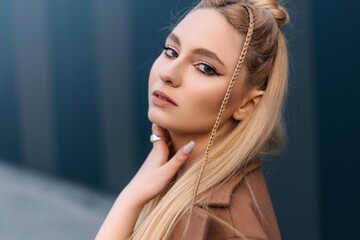 Fashionable young blonde woman with braids woven into her hair, wearing a stylish brown coat and a ring, gently touching her neck while posing in front of a modern blue building
