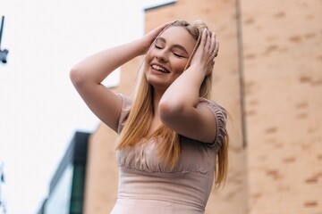 Happy fashionable blonde woman wearing trendy casual dress is touching her hair, smiling and closing her eyes while enjoying a sunny summer day outdoors in the city