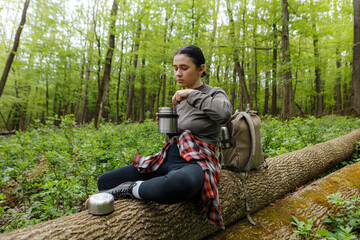 Young female hiker sitting on a large fallen tree trunk, opening her thermos to enjoy a healthy lunch, surrounded by the tranquility of a lush green forest