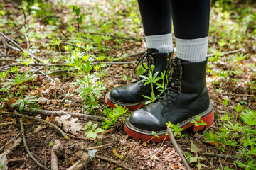 Hiker wearing black boots with red soles is standing in a wild forest, surrounded by green plants and dry branches, enjoying the peace of nature