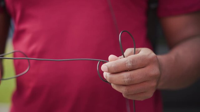 black street musician untangles worn earphones, closeup of dusty hands in red shirt manipulating tangled audio cable, careful fingers sorting cord and plug, outdoor urban sidewalk mood, preparation