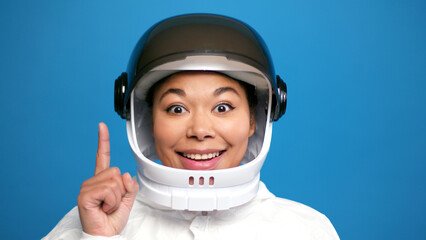 Fototapeta premium Happy hispanic woman in helmet and spacesuit pointing finger up having good idea on blue background