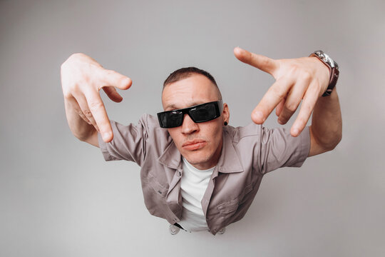 Studio portrait of a young man in gray shirt and sunglasses making expressive rapper hand gestures and facial expressions. Fisheye top-angle lens creates strong body distortion on gray background.