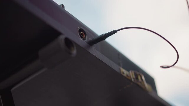 hands plugging audio cable into speaker, street DJ performing preshow setup on outdoor stage, closeup of jack insertion, warm golden light, focused fingers on analog console and rugged flightcase