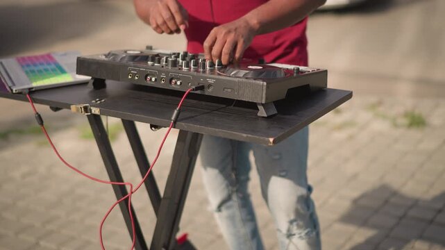 Black DJ mixing on portable controller. Sidewalk setup shows hands on mixer and pads, cables and suitcase case, sneakers and ripped jeans. Live sampling and scratching create beat drops, producer