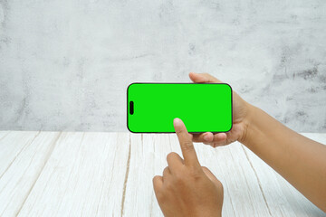 Hand holding smartphone with green screen in landscape orientation on light wall background. Concept of mobile mockup, video ads, app demo, digital marketing, and screen replacement.