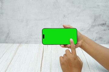 Hand holding smartphone with green screen in landscape orientation on light wall background. Concept of mobile mockup, video ads, app demo, digital marketing, and screen replacement.
