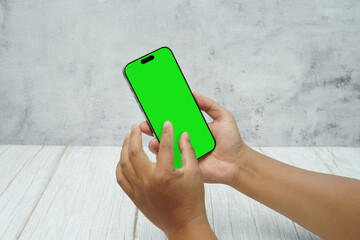 Hand holding smartphone with green screen on bright wooden desk and light wall background. Concept of mobile mockup, app presentation, digital marketing, UI UX design, and screen replacement.