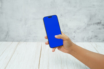 Hand holding smartphone with solid blue screen on bright wooden desk and light wall background. Concept of mobile mockup, app UI presentation, digital marketing, and modern technology.