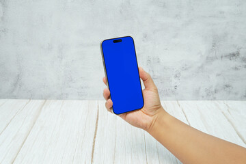 Hand holding smartphone with solid blue screen on bright wooden desk and light wall background. Concept of mobile mockup, app UI presentation, digital marketing, and modern technology.
