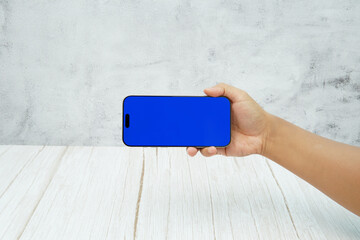 Hand holding smartphone with solid blue screen in landscape orientation on bright wooden desk and light wall background. Concept of mobile mockup, app demo, video advertising, and digital branding.