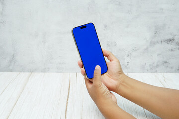 Hand holding smartphone with solid blue screen on bright wooden desk and light wall background. Concept of mobile mockup, app UI presentation, digital marketing, and modern technology.