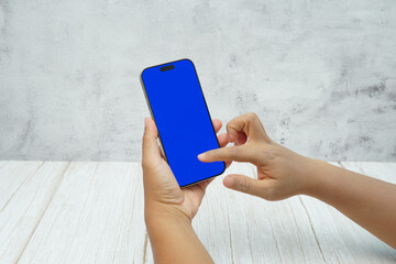 Hand holding smartphone with solid blue screen on bright wooden desk and light wall background. Concept of mobile mockup, app UI presentation, digital marketing, and modern technology.