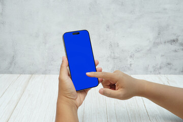 Hand holding smartphone with solid blue screen on bright wooden desk and light wall background. Concept of mobile mockup, app UI presentation, digital marketing, and modern technology.
