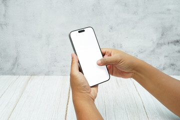 Two hands holding smartphone with blank white screen on bright wooden desk and light wall background. Concept of mobile mockup, app interaction, UI UX presentation, and digital marketing.