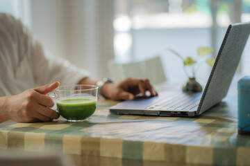 Fototapeta premium Close-up image of a male holding a glass of iced matcha green tea, sipping iced green tea and working on laptop while relaxing at a cafe.
