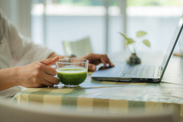 Close-up image of a male holding a glass of iced matcha green tea, sipping iced green tea and working on laptop while relaxing at a cafe.