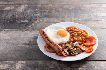 Full English breakfast with fried egg, bacon, sausages and beans on wooden table. Copy space