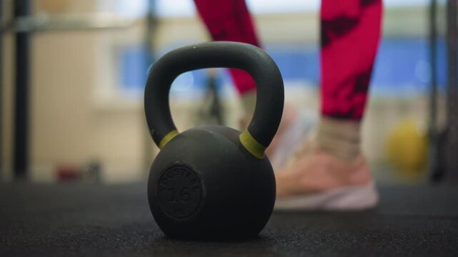 kettlebell on gym floor near athlete stepping toward handle preparing swing, bright pink leggings and sneakers, rubber flooring, blurred equipment in background, warm indoor lighting, focused training