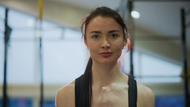 Portrait female athlete smiling in gym, postworkout glow with damp hair and light catching skin, trainer debrief and hydration break vibes, friendly gaze toward camera, pullup rig and bright window