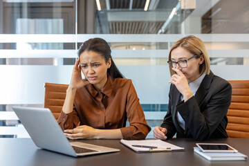 Two diverse business women in a modern office meeting room experiencing stress and confusion while analyzing a difficult problem on a laptop screen, brainstorming solutions