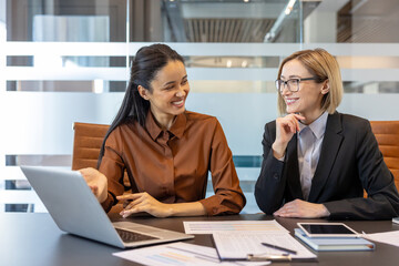 Two smiling multi-ethnic business women discussing a project, collaborating happily in a modern corporate office, using a laptop and documents to analyze data during a professional meeting