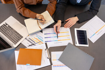 Business professionals collaborating at a desk, reviewing financial reports and spreadsheets while taking notes and using a laptop and tablet, working together on a strategic plan