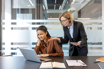 Strict female boss scolding a pressured female employee looking upset and holding her head, experiencing bullying and workplace conflict in a modern office environment
