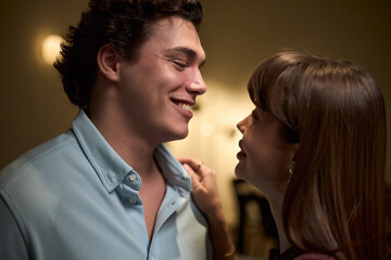 Young Caucasian man smiling while standing close to young Caucasian woman, both facing each other and sharing intimate moment indoors, soft lighting highlighting facial expressions