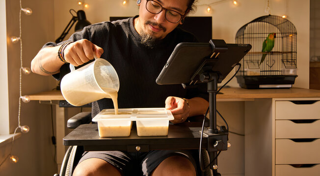 Disabled man in wheelchair pouring mixture into silicone mold for DIY soap or candle making at home with tablet and pet parrot