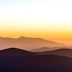 Fototapeta premium Layered mountain silhouettes fade into a warm, colorful sky at sunset or sunrise