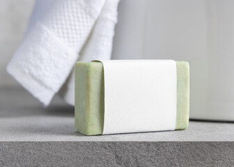 Fototapeta premium Green soap bar with blank label near white bath towels and basin on grey stone, mockup
