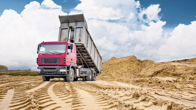 A large dump truck unloads sand at a construction site, with a large pile of sand visible in the background on a cloudy day