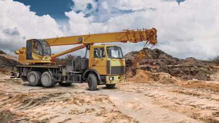 A truck crane stands ready for work at a construction site in a vast area filled with earth embankments, under a partially cloudy sky © Anoo