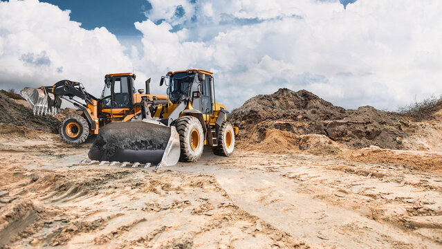 Two powerful wheel loaders stand ready to work in a sand quarry, while clouds disperse across the blue sky. The equipment is ready for construction work
